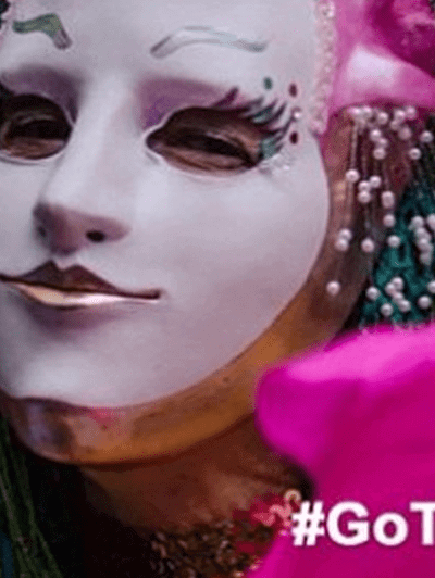 Elderly lady wearing a theatrical mask holding a hot pink fan. With #GoToWV_InstaMeet