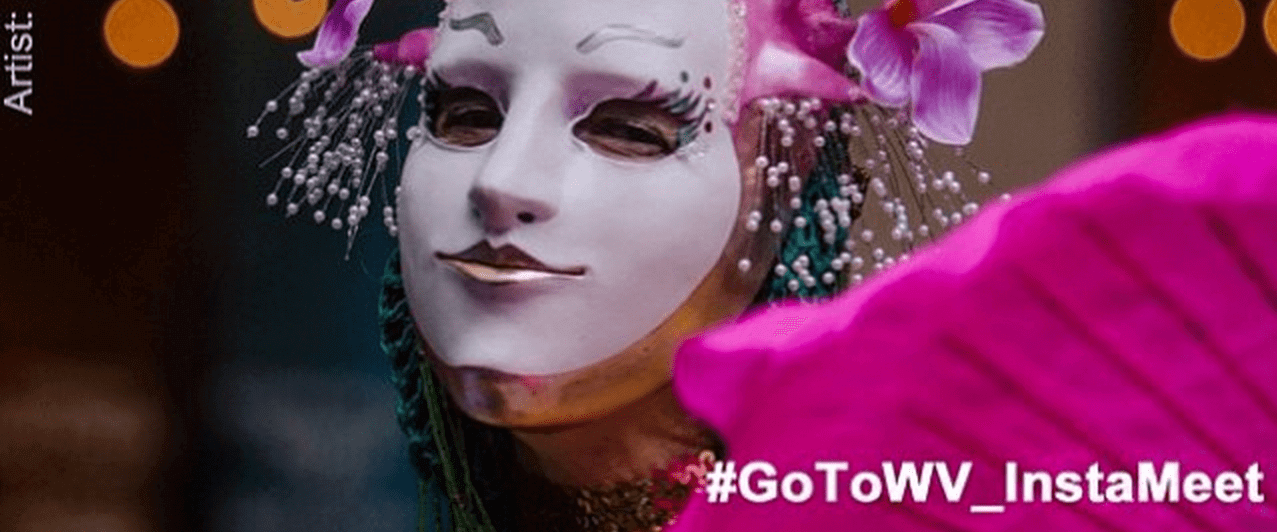 Elderly lady wearing a theatrical mask holding a hot pink fan. With #GoToWV_InstaMeet
